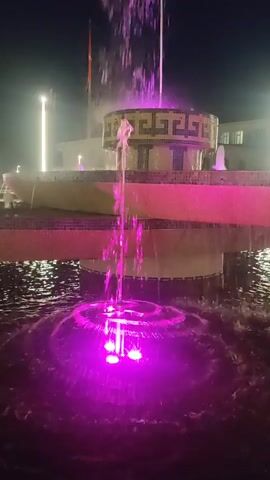 The media file depicts a nighttime scene featuring an illuminated fountain. The fountain has multiple tiers, with water spraying upwards and falling into the pool below. The water jets are lit with colorful lights, primarily in shades of pink and red, creating a vibrant and dynamic display. The fountain is situated in an outdoor setting, likely a public space given the presence of architectural elements and lighting structures in the background. These background elements include what appear to be illuminated geometric shapes and tall, modern light poles, suggesting a contemporary urban or park environment.

In the background, there are some indistinct figures that could be people, but they are not clearly discernible. The time of day is clearly night, as indicated by the dark sky and the prominent artificial lighting. The weather appears calm, with the water in the fountain flowing smoothly. The location cues, such as the architectural style of the fountain's upper tiers with its patterned rim, and the overall ambiance, align with a sense of a festive or decorative public space. The vibrant lighting and the movement of the water evoke a feeling of wonder and visual appeal, typical of illuminated fountains designed for public enjoyment. No discernible text is visible in the media file. The setting is identified as Batken, Kyrgyzstan.