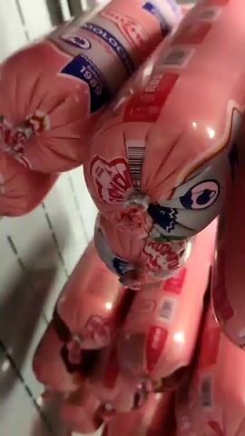 This is a close-up, vertical video of several large, pink sausages hanging in a row. The sausages are individually wrapped in plastic packaging that features pink and white branding. Some of the labels are visible, displaying text such as "ROZMETOV" and "TELYACHYA." The sausages are tied off at the ends with silver clasps. The lighting suggests an indoor environment, possibly a food production or storage area. The camera is positioned to focus on the sausages themselves, with no other objects or people clearly visible in the frame.