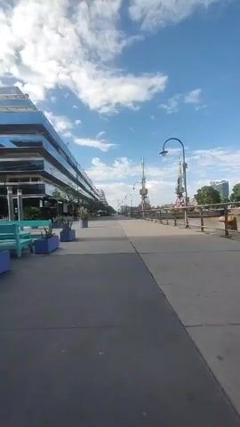 The video shows a view of a waterside promenade or pier on a bright, sunny day. On the left is a modern building with multiple floors and a blue mirrored facade. Alongside the building are planters containing greenery and turquoise benches. The promenade stretches into the distance, flanked on the right by a railing and the water's edge. In the middle ground, there are several large cranes, remnants of the port's industrial past. The sky is blue with scattered white clouds, and the sun is visible. The scene is quiet and appears to be devoid of people.
