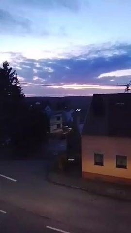 The video shows a residential street corner at dusk. The sky is a mix of blue, gray, and purple hues, with a line of pink on the horizon. On the left side of the frame is a tall evergreen tree, partially obscuring a building behind it. To the right, a two-story building with a lighted window is visible. A road with white lane markings runs along the bottom of the frame, curving into the distance. A white car is seen making a right turn onto the road. Soon after, a dark-colored car drives by in the same direction. The camera appears to be stationary, possibly positioned from a higher vantage point, like a balcony or upper-floor window.