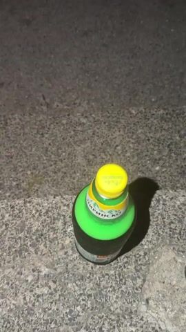 A green glass bottle with a yellow cap sits on a textured gray surface, possibly concrete steps or pavement. The bottle appears to contain a green liquid. The lighting suggests it is nighttime or a dimly lit indoor setting. The bottle has visible text on its label, though it is partially obscured and difficult to read.