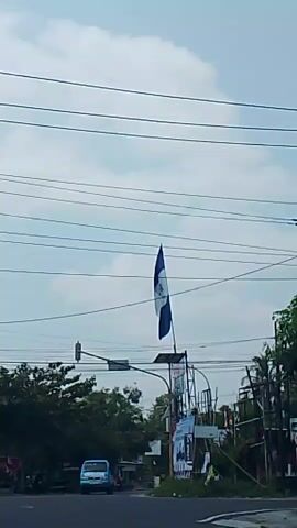 The video shows a street scene with a blue and white flag prominently displayed on a pole in the center. The flag appears to be the flag of El Salvador. There are power lines overhead and a traffic light in the background. On the street, a blue pickup truck is driving towards the camera, and a yellow truck is also visible further down the road. There are trees and buildings on either side of the street, and the sky is cloudy. Some posters or banners are visible on a structure to the right of the flag pole. The overall impression is of a street in a town or city.