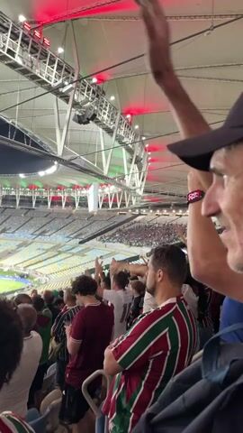 This video shows a crowd of people attending a sporting event. The camera is positioned among the spectators, looking towards the field and the opposite stands. Red and green lights are visible illuminating the stadium's ceiling. Many people in the foreground are wearing shirts with green and maroon stripes, suggesting they are fans of a particular team. Some individuals are raising their hands, cheering or waving. The field of play is visible in the background, with players on the grass and more spectators in the distant stands. The atmosphere appears energetic and celebratory.