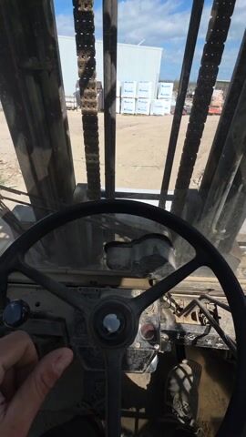The video captures the view from inside a forklift operating in an outdoor environment. The camera is positioned behind the steering wheel, which is the primary focus in the lower portion of the frame.  The forklift's chains and vertical support structure are prominently visible through the front windshield. The outside environment is sunny with some clouds in the sky. A large pile of discarded materials, including wood scraps and pallets, is visible in the middle ground. In the background, there are other vehicles and structures, including a truck and a large warehouse. A hand briefly appears above the steering wheel towards the start of the video.