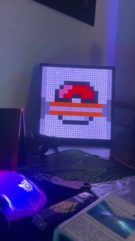 The camera is positioned at a medium distance, slightly angled down, providing a view of a gaming desk setup. A small pixel art display screen is the focal point, showing various blocky character faces that change color. The screen rests on the desk against a wall. In the foreground, a purple glowing Razer mouse is visible on the left side. A green Razer mousepad, a lighter, and a pack of what appears to be cigarettes are also on the desk. Behind the pixel art screen, there is a speaker. The room is dimly lit with a blue-purple hue dominating the scene.