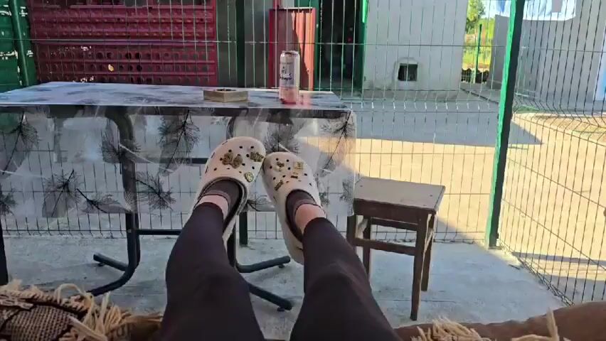 This is a first-person perspective video taken outdoors on a sunny day. The viewpoint is from someone lying down, with their legs and feet propped up on a surface, possibly a sofa or lounger. Their feet are clad in white Crocs adorned with several charms, including what appears to be a bee or butterfly, and possibly a smiley face. The person is wearing black leggings. In the background, there is a table covered with a transparent plastic sheet featuring a black leaf pattern. On the table sits a silver can, possibly a beverage, and an ashtray. A small wooden stool is visible to the right of the table. The area appears to be a patio or porch, with a concrete floor and a fence surrounding the space. Beyond the fence, some structures and greenery are visible. The camera occasionally pans slightly, showing the environment and the person's own legs and feet.
