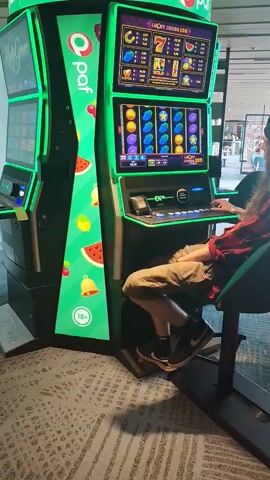 A person with long brown hair and a red plaid shirt is sitting in a black chair in front of a green and black slot machine. They are wearing khaki shorts and black sneakers with white socks. The slot machine has a screen displaying colorful symbols, including cherries, watermelons, and bells. The person's right hand is on the control panel of the slot machine. To the left of the person is another slot machine with a similar design. The floor is covered with a patterned carpet. In the background, there are other slot machines and a few people walking by. The overall environment appears to be a casino or gaming area.