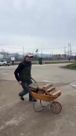 A man pushes a yellow wheelbarrow full of split logs across a concrete surface toward an open area with flags, lighting towers, and a building in the background. The man wears a tan baseball cap, a dark vest over a long-sleeved shirt, jeans, and dark sneakers. He smiles and struggles slightly with the wheelbarrow. The sky is overcast.