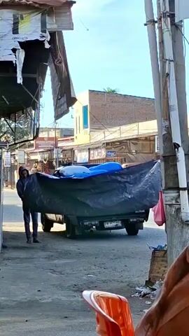 A person wearing a hooded jacket walks on the left side of the frame, holding a large black tarp that is partially covering the bed of a pickup truck. The truck is parked on a dirt road and appears to be loaded with blue objects covered by the tarp. In the background, there are buildings and a clear sky. A motorcycle is visible passing by in the distance. The scene appears to be an outdoor market or street.
