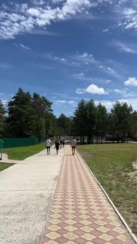 A vertical shot shows a group of people walking on a paved path on a sunny day. The path is divided into two sections: a wider concrete section on the left and a narrower section with a red and yellow patterned tile on the right. Several trees line the right side of the path, and a green fence is visible on the left. The sky is blue with scattered white clouds. Six people are walking away from the camera on the path. They are all wearing casual clothing. The person in the foreground on the right is wearing a white t-shirt and black shorts. To their left and slightly ahead, two people are walking together, one in a white t-shirt and black shorts, and the other in a black t-shirt and black shorts. Further ahead, another group of three people are walking. The person in the middle is wearing a white t-shirt and dark shorts, and appears to be holding something red. The person behind them is wearing a light-colored t-shirt and dark shorts. The person in front of them is wearing a blue t-shirt and dark shorts. The overall impression is a leisurely walk in an outdoor setting.