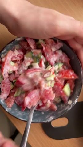 A close-up shot shows two hands mixing a salad in a dark blue bowl with a spoon. The salad is composed of chopped red tomatoes, sliced green cucumbers, and what appears to be chopped onions and dill, all coated in a creamy white dressing. The hands are gently stirring the ingredients, moving the spoon through the salad to combine them. The bowl sits on a wooden surface, likely a table. The lighting suggests an indoor setting. The video is short and focuses on the action of mixing the salad.