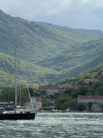 A wide shot captures a bay with calm water in the foreground, reflecting the cloudy sky and lush green mountains in the background. A white sailboat with a tall mast is docked to the left. Along the shore, nestled amongst trees, are several buildings with red tile roofs, characteristic of Mediterranean architecture. The mountains rise steeply behind the village, covered in dense vegetation, with a few patches of exposed rock. The overall atmosphere is serene and picturesque.