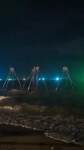 The video shows a nighttime beach scene with the ocean in the foreground and three strange structures standing in the water. The structures have a central body and three long, thin legs, giving them an alien or otherworldly appearance. They are illuminated by lights from the background, creating a surreal atmosphere. The ocean waves are visible, gently washing onto the shore. The background lights are blurred, suggesting they are distant city lights or boats. The sky is dark, adding to the mysterious feel of the scene.