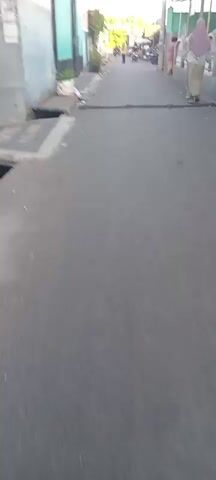 The video shows a narrow asphalt street. There are buildings on both sides of the street, with some greenery visible on the left. A person wearing light-colored clothing is walking away from the camera on the right side of the street. The overall environment appears to be an outdoor, urban or semi-urban setting.