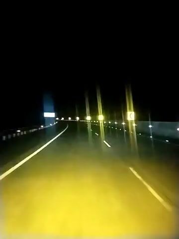 The video shows a first-person perspective of driving on a highway at night. The road is illuminated by the headlights of the vehicle and by streetlights or reflective markers lining the highway. The sky is completely dark. The highway appears to have at least two lanes in the direction of travel. Road signs, some illuminated and some reflective, are visible along the side of the road, indicating curves and direction. The overall impression is of a deserted highway with good visibility due to artificial lighting.