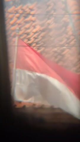 This is a very blurry, close-up video of an Indonesian flag. The flag is red and white and appears to be attached to a flagpole. The background is also out of focus, but it looks like a tiled roof. The video is very short and appears to be handheld. There are no people or other objects clearly visible in the frame.