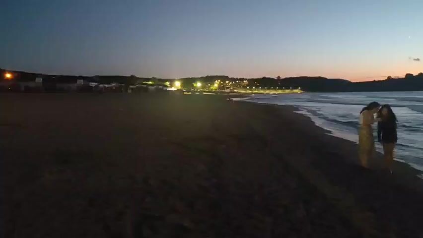 Two people are on a sandy beach at dusk. The person on the left is wearing a patterned dress and the person on the right is wearing a dark jacket and pants. They are standing close together near the water's edge. The ocean is to their right, with waves gently rolling onto the shore. In the distance, a town or village is visible with lights illuminating the buildings and a road or promenade. The sky above is a deep blue, fading to a lighter orange and pink hue near the horizon, indicating either sunrise or sunset. The overall atmosphere is calm and serene.