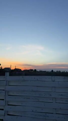 The video shows a plane flying over a small town during sunset. In the foreground, there's a white wooden fence. Beyond the fence are houses, power lines, and trees silhouetted against the colorful sky. The sky blends from light blue at the top to orange and pink near the horizon, where the sun is setting. The plane is a dark silhouette against this vibrant sky. There are also a few birds flying in the distance.