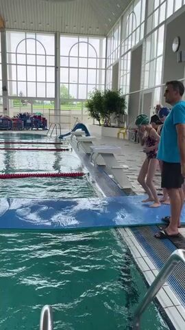 A child in a swimsuit and swim cap is seen running across a partially submerged blue mat in an indoor swimming pool. The child is barefoot and moving quickly. The pool is surrounded by large windows and there are two adults standing nearby observing.  The interior of the building has high ceilings and wooden beams. There is a tree and exercise equipment nearby.
