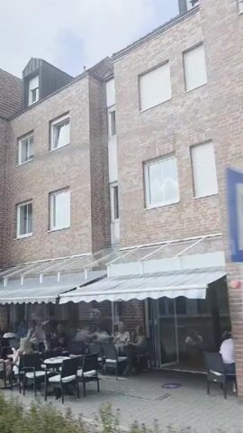 The video pans across a street scene featuring a multi-story brick building with several windows and an awning covering an outdoor seating area where people are seated at tables. A sign for "SCHMITJANS Immobilien" is visible. To the left, another building with a yellow sign that has an ice cream cone graphic is partially visible. A woman on a bicycle rides past the building. Further down the street, another building has signs for "Immobilien" and "LANGER." In the background, there are parked cars, bicycles, and more buildings with houses visible. The sky is partly cloudy.