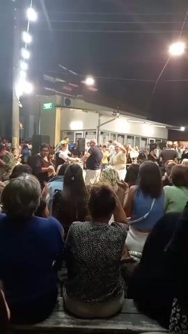 A crowd of people is gathered outdoors at night, watching a performance. Strings of lights are visible overhead, and a building with windows and a sign is in the background. Several individuals are dancing and performing in front of the audience. The audience members are seated, facing the performance area. Some individuals in the crowd are holding up their phones, possibly recording the event. The atmosphere appears to be festive.