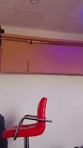 A stationary shot focuses on an interior space with a low-angle perspective. A red barstool with chrome arms and footrest is positioned in the foreground. The background consists of a white wall, and a suspended roller blind in a light brown color is hanging close to the ceiling. Two light sources are present on the ceiling: a recessed can light with a white cover and a blue LED light. A dark object partially enters the frame from the left. The overall lighting creates a somewhat sterile and contemporary aesthetic.