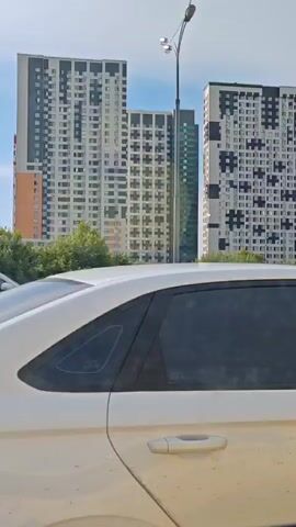 The media file shows a view from inside a car looking out the passenger side window. The car is white and appears to be driving on a highway. In the background, a line of tall, modern apartment buildings is visible. These buildings have varied architectural styles and colors, with some featuring black and white geometric patterns and others being solid colored. Green trees and foliage line the area in front of the buildings. The sky is blue with some scattered clouds, suggesting a clear, sunny day.

The activity depicted is driving on a highway, indicated by the movement implied by the camera's perspective and the presence of other vehicles on the road. A dark gray SUV is visible in the right lane, and other cars can be seen further in the distance. One of the apartment buildings has text visible on its facade that appears to say "самолет". The time of day seems to be daytime, likely late morning or afternoon, given the bright sunlight and shadows. The scene is urban or suburban, with a focus on modern residential architecture. No specific emotions are conveyed, but the setting suggests a typical day in a modern city or its outskirts. The location context provided is Razdory, Russia, which aligns with the visual cues of the architecture and the Russian text.