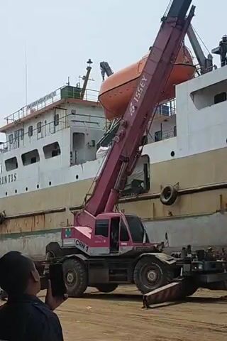 This media file captures an industrial scene, likely at a dock or shipyard in Kamal, Indonesia. The primary focus is a large, white and yellow ship, appearing to be a passenger ferry, docked alongside a sandy or dirt lot. A prominent feature is a large, pink crane, identified with the text "ISSEY" and "CRN 1," actively engaged in an operation.

The crane is lifting a life raft, a cylindrical white object with orange ends, from the ship. The life raft is suspended by cables and is being lowered towards a red flatbed truck parked beside the ship. A person in a white hat is visible on the truck, appearing to direct or prepare to receive the life raft.

On the deck of the ship, several more life rafts are visible, some neatly stacked. A few individuals are also present on the ship's deck, observing the operation. The overall atmosphere suggests a practical, work-oriented environment.

The lighting indicates it is daytime, with bright, diffused sunlight, suggesting clear weather. The scene is set against a pale blue sky. The ship itself shows signs of wear, with rust and grime visible on its hull, particularly along the waterline and the lower yellow section, indicating it might be undergoing maintenance, repairs, or decommissioning. The sandy ground and the presence of heavy machinery like the crane and truck further reinforce the industrial setting. The activity depicted is the removal or transfer of equipment from the ship.