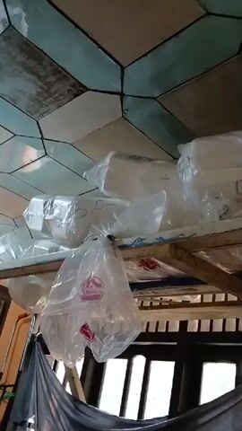 The video shows a ceiling covered with geometric tiles in shades of beige and teal, some of which appear weathered. Below the ceiling, a wooden shelf holds several white objects wrapped in clear plastic bags. Some of these bags are tied shut, while others are open. One plastic bag hangs precariously from the shelf, obscuring a portion of the items behind it. Further down, a dark tarp is draped over some wooden beams, and behind it, a dark doorway or window is visible with light filtering through. A glimpse of an orange object, possibly a broom handle, can be seen on the left side of the frame. The overall impression is one of storage in a somewhat cluttered or unfinished space.