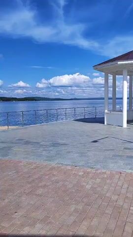 The video shows a view of a body of water, likely a lake or sea, under a bright blue sky with scattered white clouds. In the foreground, there is a paved walkway with a red brick border. Beyond the walkway is a grey stone paved area, and further still is a metal railing along the edge of the water. To the right of the frame, there is a white gazebo with a dark roof and supporting pillars. The gazebo is situated on the paved area near the railing. The water extends to the horizon, where a tree-covered shoreline is visible in the distance. The overall scene depicts a pleasant, outdoor, waterfront location on a clear day.