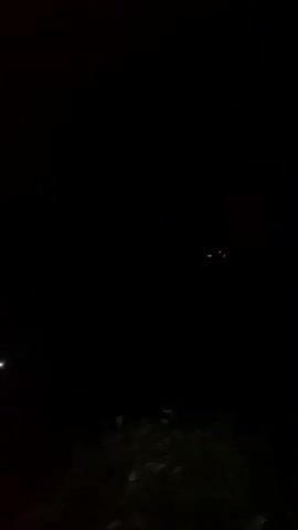 This video shows a nighttime scene with a dark sky. In the upper central part of the frame, a small, airborne object with several lights is visible. The object appears to be a drone or a similar flying device, with multiple small lights that are white or bluish, and possibly a red light as well. The object is not clearly defined due to the darkness and distance, but its shape suggests it could be a quadcopter or a similar multi-rotor drone. In the lower left portion of the frame, there is a faint, diffused light source, possibly a distant streetlamp or porch light, casting a dim glow on a shadowy structure. The lower portion of the frame also shows dark foliage, likely bushes or trees. The overall impression is a recording of a drone flying in a dark, outdoor environment.