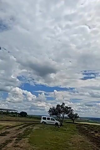 A white van is parked on a grassy, uneven terrain. Two trees stand near the van, casting shadows. The sky is filled with dramatic, billowy clouds, with patches of blue visible. The background shows rolling hills and fields, suggesting a rural or countryside setting. The camera is angled upwards, focusing on the sky and the surrounding landscape.