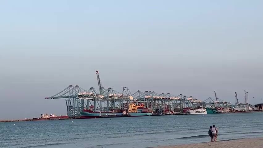 The video shows a wide shot of a busy port with many large shipping cranes and several cargo ships docked. The water in the foreground is calm, with gentle waves lapping at a sandy beach. Two people are walking along the shoreline in the distance. The sky is clear and light blue, suggesting it is either early morning or late afternoon. The overall impression is one of industrial activity juxtaposed with a tranquil beach scene.