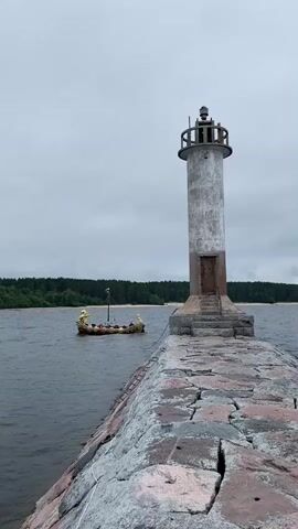 The video opens with a view of a lighthouse on a stone pier, with a Viking-style boat in the water to the left. The sky is overcast. As the camera pans to the right, the lighthouse recedes and a treeline comes into view along the distant shore. Further panning right reveals a beach, more trees, and eventually a building with a unique architectural style on the left side of the frame. The boat remains visible for the first few seconds of the pan. The water is calm throughout.