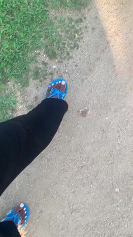 This is a first-person perspective video of someone walking on a dirt path next to a grassy area. The person is wearing black pants and blue flip-flops with a white bow detail. Their toenails are painted white. The scene appears to be outdoors in a sunny environment, as evidenced by the shadows cast on the ground. The overall impression is one of a casual walk in a natural or semi-natural setting, possibly in Reak, Nigeria. There is no visible text in the video. The time of day appears to be daytime, and the weather seems pleasant enough for walking outdoors.