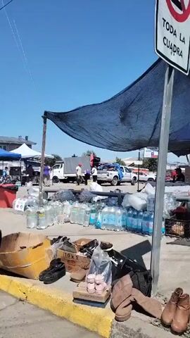 The video shows a parking lot that has been converted into a donation distribution center. There are several white and blue canopies set up in the parking lot, and people are milling around them. There are also piles of water bottles, boxes of shoes, and other items stacked on the ground. There is a sign that says "Comida Gratuita" which translates to "Free Food". There are also several vehicles parked nearby, including trucks and cars. The sky is blue and clear.