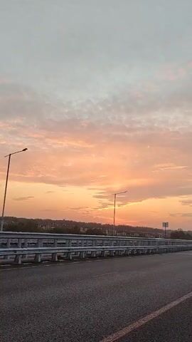 The video shows a time-lapse of a sunset with a dramatic sky filled with orange and pink clouds. A highway bridge railing is visible in the foreground, with a road stretching out ahead. Streetlights are positioned along the railing. In the distance, houses and trees line a gentle slope. The sun is setting behind the horizon, casting a warm glow over the scene.