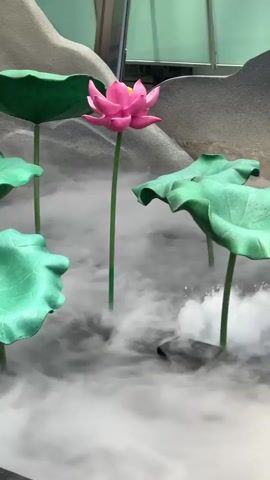 The video shows a display of artificial lotus flowers and leaves amidst a cloud of fog or vapor. The flowers are pink, while the leaves are green, and are positioned at varying heights. A backdrop resembling a gray mountain range is visible in the background.  The fog creates an ethereal effect, partially obscuring the base of the plants.  The scene appears to be indoors, possibly in a shopping mall or an event space.