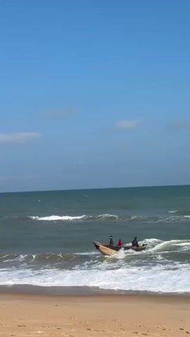 The video shows a bright, sunny day at a beach, likely in Badagry, Nigeria. The sky is a clear, vibrant blue with a few scattered white clouds. The ocean is a deep green, with moderate waves breaking near the shore, creating white foam and spray. The sand on the beach is a light brown.

The central focus is a wooden boat with several people in it. The boat is partially in the water and partially being pulled towards the shore by a group of men on the beach. The people in the boat appear to be a mix of adults and possibly older children, dressed in casual clothing. Some are actively engaged in maneuvering the boat, while others appear to be passengers.

On the beach, several men are actively involved in helping to bring the boat in. They are pulling ropes attached to the boat and appear to be working together to overcome the resistance of the waves and sand. Their movements suggest effort and coordination. The overall atmosphere seems to be one of communal activity and perhaps a routine task of beaching a fishing boat or similar vessel.

The time of day is clearly indicated by the bright sunlight and the quality of the shadows, suggesting it is likely late morning or early afternoon. The weather is warm and pleasant, with no indication of rain or strong winds, beyond the natural movement of the ocean waves. There is no visible text in the video. The scene is evocative of coastal life and the interaction between people and the sea.