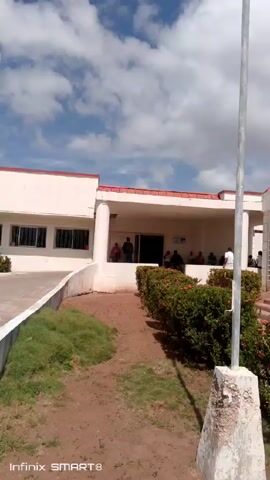 The video shows an exterior view of Clinica Popular El Espinal. The building is a single-story structure with a white facade and a red roof. The sign above the entrance reads "Clinica Popular El Espinal" in blue letters. There are several people visible in front of the clinic, and cars parked in the parking lot. The sky is blue with scattered white clouds. The ground is paved with a concrete walkway and a grassy area with bushes.