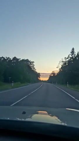 The video shows a first-person perspective of a car driving on a two-lane asphalt road through a forested area at dusk. The sky is clear with a gradient of light blue to pale orange near the horizon, indicating the sun is setting. Trees line both sides of the road. The road has white lane markings. In the foreground, the dashboard and windshield of the car are visible, with a reflection of the sky and trees on the windshield. A speed limit sign of 70 is visible on the road.