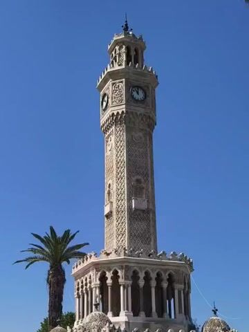 The video shows a tall, ornate clock tower made of light-colored stone, possibly marble. The tower has multiple tiers and is decorated with intricate carvings and what appear to be Islamic-inspired motifs. A clock face is visible on one side of the tower. At the base of the tower, there is a hexagonal structure with arches and decorative elements. To the left of the tower, a tall palm tree stands against a clear blue sky. The ground around the tower appears to be paved. Further into the background, other palm trees and greenery are visible, along with some people walking and benches. The lighting suggests it is daytime. The camera pans slightly to the right, showing more of the plaza area, with more palm trees and paved pathways. Pigeons are visible on the ground in the foreground.