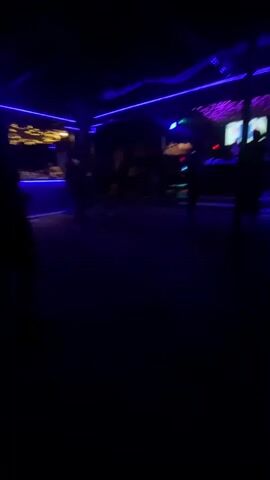 The video captures a dimly lit night club scene with blue and purple ambient lighting. Silhouetted figures are scattered across the dance floor. In the background, a DJ booth is visible, illuminated with a pink and white display. Other lights flash, contributing to the atmosphere. A few individuals are standing near the DJ booth.
