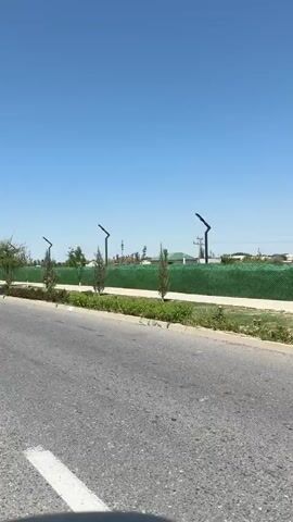 The video displays a bright, sunny day with a clear blue sky, suggesting it's daytime. The scene is captured from a moving vehicle, looking out towards a landscaped area adjacent to a road. The road itself is paved and appears to be a street or highway, with a white line marking the lane.

On the left side of the road, there's a meticulously maintained green space. This area features a variety of trees, some tall and slender like cypress trees, and others with fuller canopies. Small, decorative plants and flower beds are visible, adding color and texture to the landscape. A low, dark-colored border separates the grassy area from a colorful flower bed.

Beyond the greenery, a tall, dense, green fence stretches across the frame. This fence likely encloses a property or park. Several modern, black lampposts with angled, light-emitting tops are spaced along the fence line, suggesting the area is designed for nighttime illumination. In the background, beyond the fence, hints of buildings and rooftops can be seen, along with more trees and structures. The architecture suggests a residential or possibly a communal area.

The overall impression is one of a well-developed and aesthetically pleasing urban or suburban environment in what appears to be a warm climate. The lack of people or specific activities points towards a general view of the surroundings rather than a focus on an event. The location is identified as Pakhtakor, Uzbekistan. No visible text is present in the video.