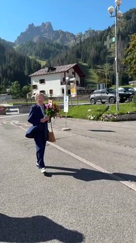 The video shows an elderly person wearing a navy blue suit and white shoes, walking with a bouquet of flowers and a brown bag. The person is walking on an asphalt surface, possibly a road or parking lot. In the background, there are mountains, trees, and buildings, including a white house with a red roof, a wooden building, and a building with flower boxes on the balconies. Several cars are parked in the background, including a silver SUV, a red SUV, and a white car. There are also some traffic cones and a street light visible. The weather appears to be sunny.