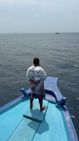 The video shows a man standing on the bow of a blue and white boat, facing away from the camera and towards the ocean. He is wearing a white hooded sweatshirt with the words "Go well Together" printed on the back, along with a picture of a whale tail, and dark patterned shorts. He is barefoot. The ocean is calm and dark blue, and there are two boats visible in the distance. The sky is overcast. The boat is moving through the water. The man takes a phone up to his ear.