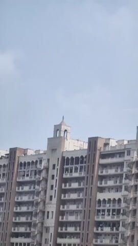 The video shows a tall apartment building with multiple stories. The building has a beige and white facade with many windows and balconies. There are spiral staircases on the exterior of the building. The sky is cloudy and overcast. The video is filmed from a low angle looking up at the building. There are no people visible in the video.