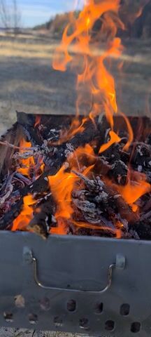 Flames engulf a pile of wood inside a metal barbecue grill. The fire emits intense orange flames and light. The metal grill has a handle on its right side. The background is blurry, with hints of grass and trees visible. The video is shot from a close-up perspective, focusing on the burning wood.