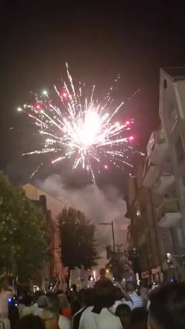 A nighttime view of a fireworks display above a street lined with buildings. A crowd of people are visible in the foreground, looking up at the fireworks. The fireworks are bursting in a variety of colors, including white, pink, and red, and are creating trails of smoke in the dark sky. Some of the buildings have balconies. Trees are also visible along the street. Several people in the crowd are holding up their phones to record the event.
