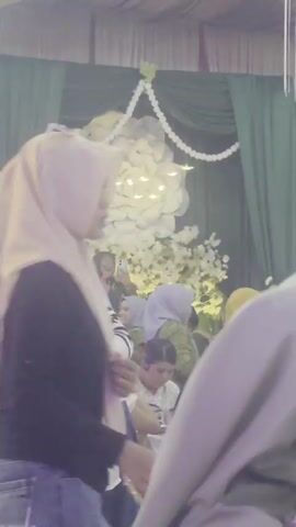 The video shows a gathering of people, likely at an event. In the foreground, two individuals wearing hijabs are partially visible, one in a purple hijab and the other in a grey hijab. Behind them, more people can be seen, including a woman in a purple hijab and glasses who is smiling. The background features a decorative wall with large white floral arrangements and green drapery. The lighting appears to be soft and ambient, suggesting an indoor setting.